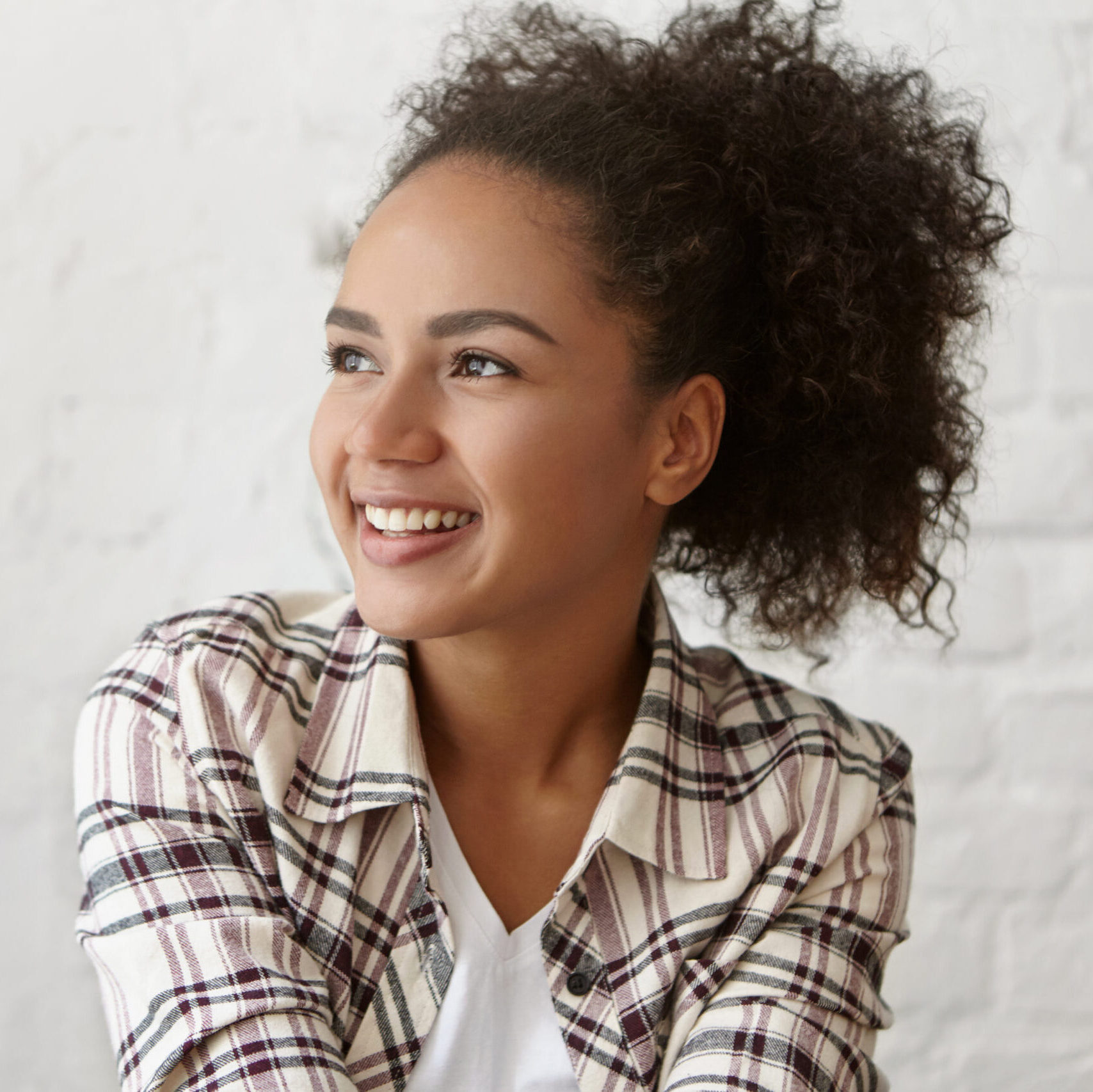 Mixed race female with Afro hairstyle wearing casual shirt looking with smile aside having dreamy expression posing against white brick wall. Restful woman with curly hair having pleasing thoughts