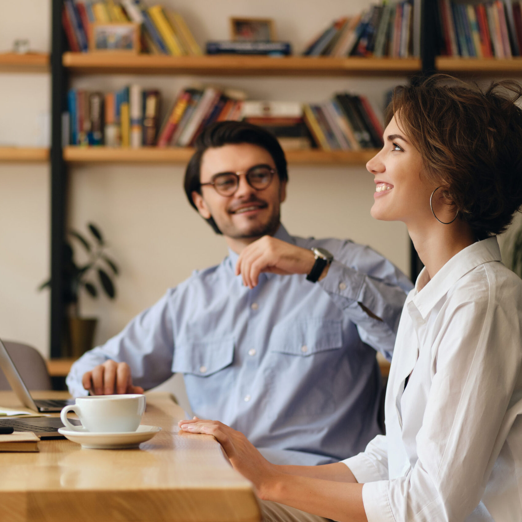 Young attractive business colleagues sitting at the table with laptop and coffee happily talking at work in modern office